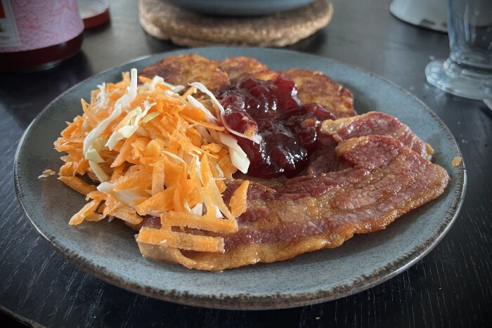 Stekfläsk och potatisbullar med vitkål- och morotssallad samt lingonsylt på en blå tallrik.