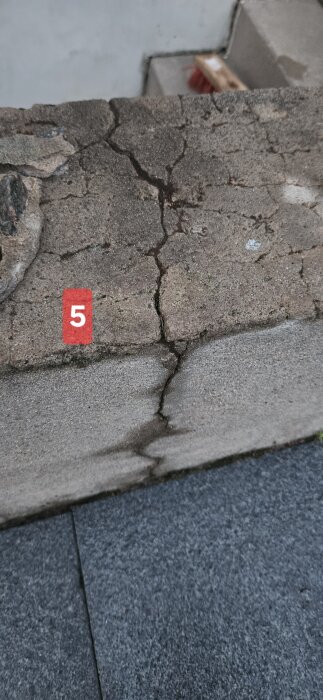 Sprickbildning i betongvägg markerad med rött nummer 5, kanske på grund av slitage. Bakgrund en del av en trappa och en borste.