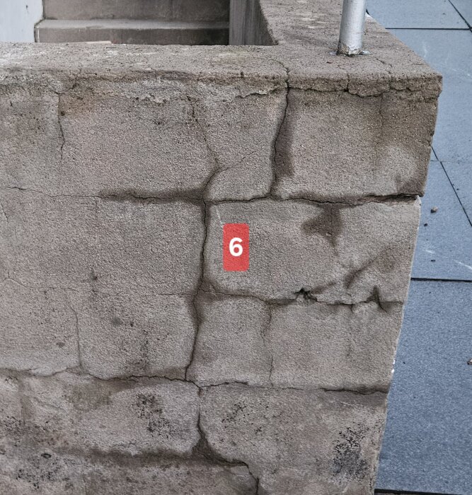 Sprucken betongvägg med numret 6 i rött. Synliga sprickor och skador i muren, eventuellt av lecablock. Trappsteg synliga i bakgrunden.