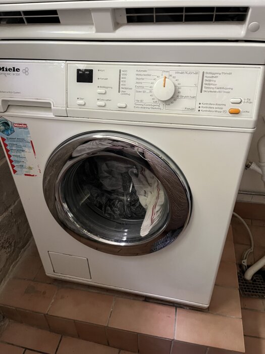 Vit tvättmaskin av märket Miele, placerad på ett klinkergolv. Maskinens frontvred och lucka syns tydligt.