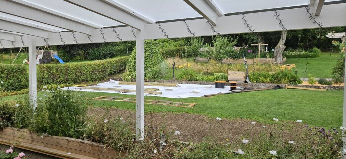 Byggarbetsplats i trädgård med träreglar och betongplattor under ett vitt tak, omgiven av grönska och blomsterplanteringar.