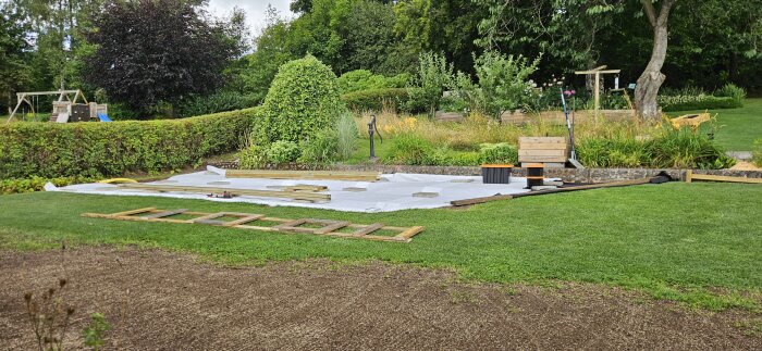 Trädgård med pågående byggprojekt för ett pooldäck. Regelverk och plattor är utlagda på marken framför häckar och träd.