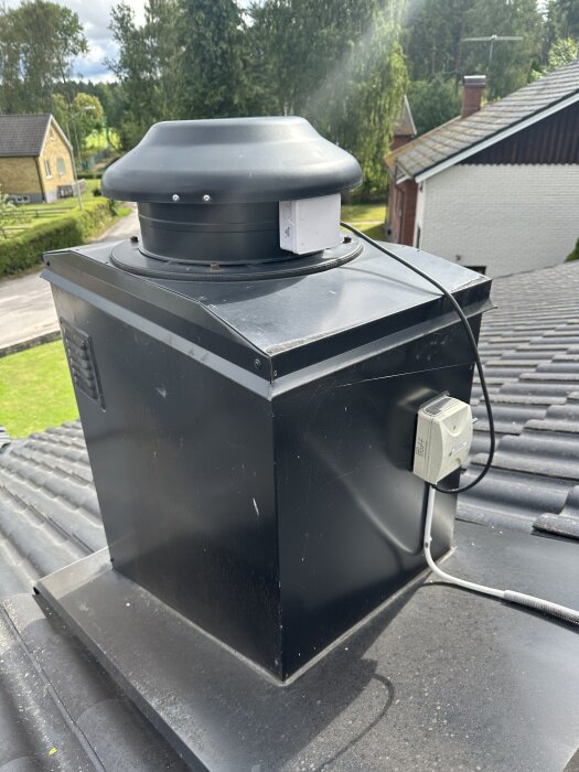 Svart takfläkt installerad på ett tak med elinstallation synlig. Skog och hus i bakgrunden under en mulen dag.