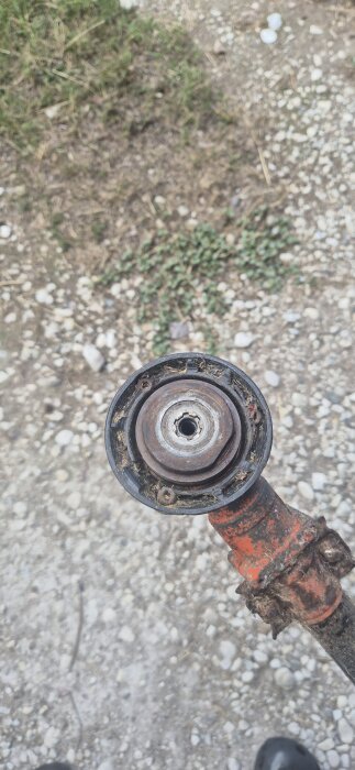 Närbild av vinkelväxel och monteringsdetaljer på en röjsåg, med gräs och stenar i bakgrunden.