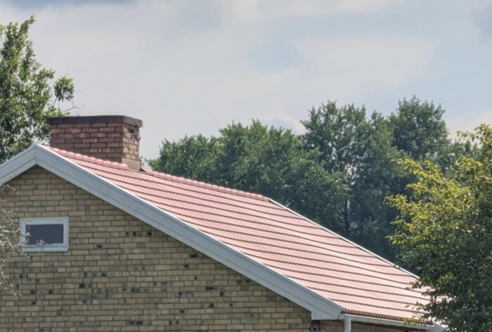 Tegeltak på en tegelbyggnad med en skorsten, omgiven av träd och en blå himmel i bakgrunden.