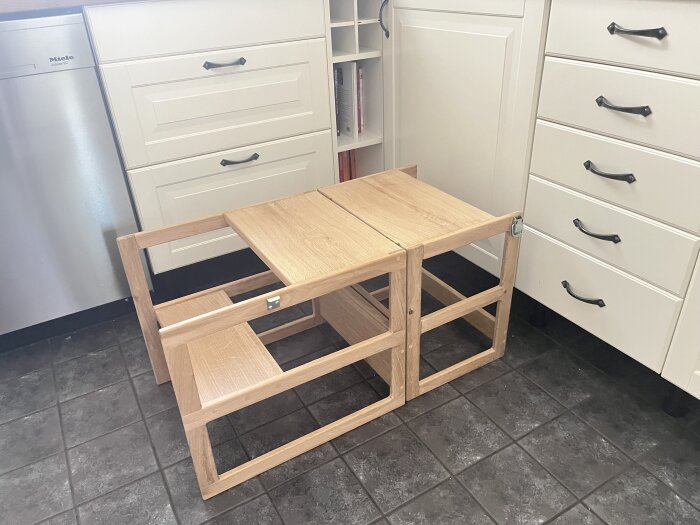 En enkel träpall/ritbord i ljus träfärg står på ett klinkergolv i ett kök, omgiven av vita skåp och en diskmaskin.