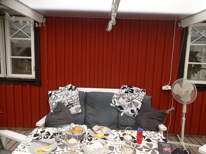 Altan med kanalplasttak, röd trävägg, ljusslinga, sittplats med kuddar och filt, matbord med mat, fläkt på golvet, fönster med korspröjs.