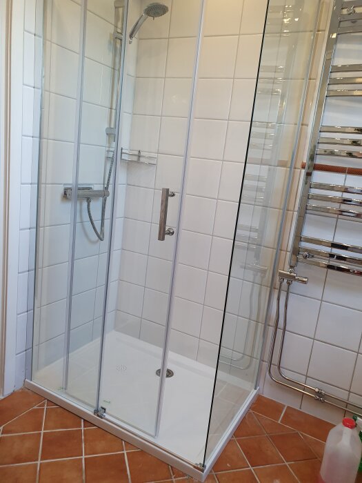Duschkabin med vita kakelväggar och genomskinliga glasdörrar, stående på ett vitt duschkar över rödbrunt klinkergolv.