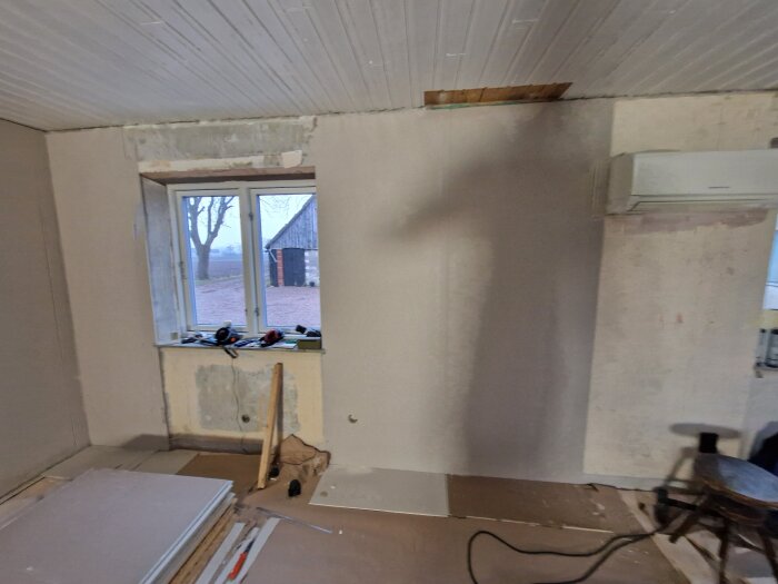 Rum under renovering med uppsatta gipsskivor, fönster som vätter mot gården, verktyg på fönsterbrädan, synliga mögelskador nära golvet.