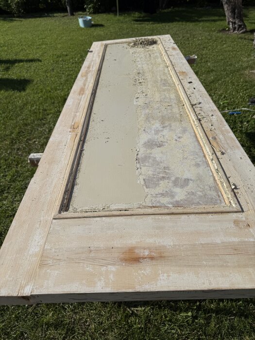 Gammal dörr under renovering försedd på gräs, med bortskrapad färg och trädetaljer blottade, en del av ett pågående renoveringsprojekt.