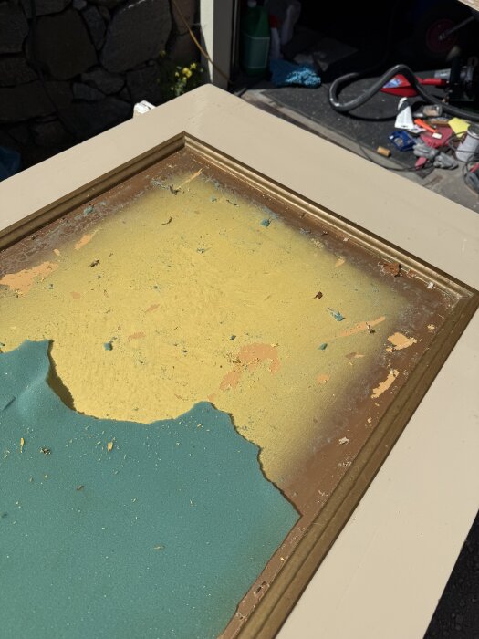 Gammal dörr renoveras med färg bortskrapad i lager visar gul och grön färg, omgiven av verktyg och material i bakgrunden.