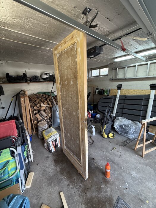 Gammal dörr som renoveras i garage, omgiven av verktyg och material. Dörren hängs upp av en tving från taket. Ved och trädgårdsutrustning i bakgrunden.