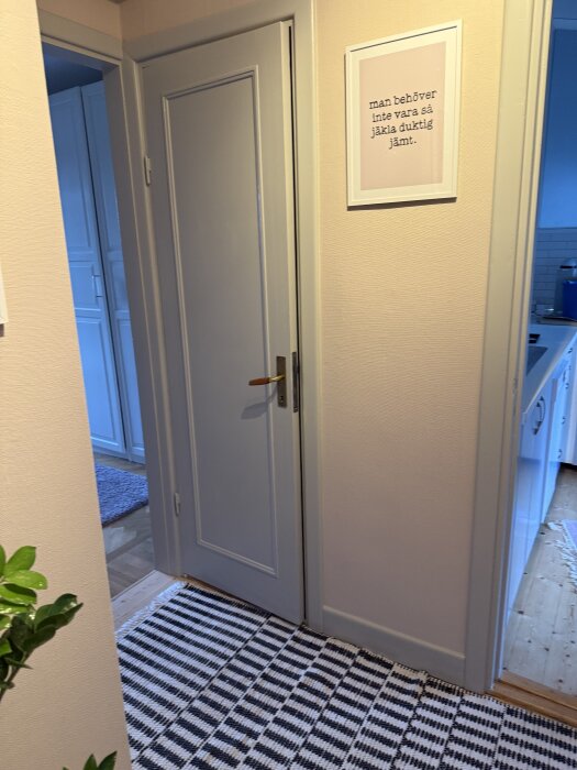Nymålad dörr i inre hall med citattavla ovanför. Svartvit randig matta på golvet och ett skymtande kök i bakgrunden.