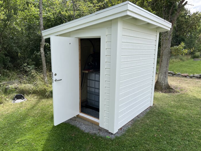 Isolerat vitt pumphus med dörr öppen, visar en IBC-tank och pumputrustning på gruset inuti, omgiven av grönskande trädgård.