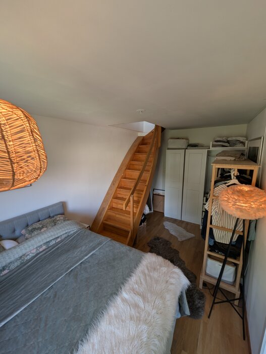 Trätrappa i ett sovrum, intill en vägg med garderober och kläder, med en säng och dekorativ belysning i rummet.