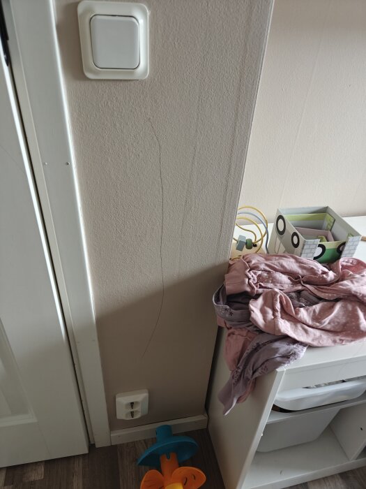 Vägg med blyertsstreck intill dörrkarm, klädhög på en vit hylla, strömbrytare och eluttag synliga.