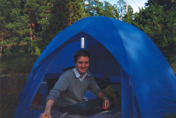 Man sitter i ett blått tält ute i naturen, ser glad ut.