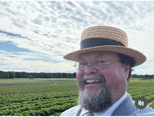 En man i halmhatt ler stort framför en fält med grödor under en molnig himmel.
