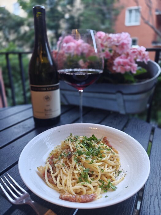 Tallrik med pasta och örter, flaska rödvin, vinglas och blommor i en kruka på ett bord utomhus.