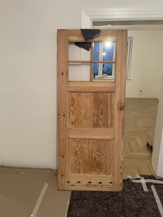 Rustik innedörr i trä lutad mot en vägg, med tre glasöppningar upptill. Dörren står i ett rum med ljusa väggar och parkettgolv.