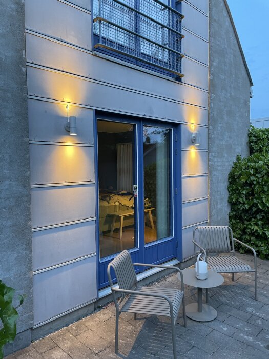 Tegelhus med blå entrédörr och två utelampor. Två stolar och ett bord står utanför. Växtlighet syns till höger. Sen kvällsbelysning.