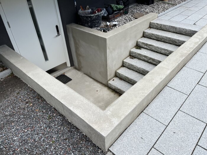 Stödmur i murad leca, putsad med cement, bredvid en entré, med tillhörande trappa av stenplattor och stenbelagd mark.