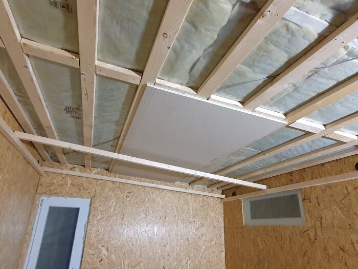 Trätak med glespanel och isolering syns ovanför ett rum med monterade gipsskivor, där en del av gipsen är på plats.