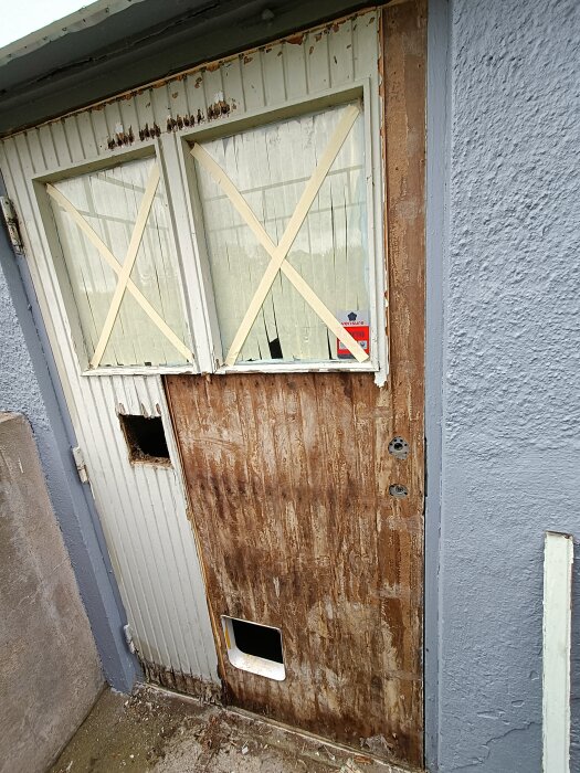 Sliten källardörr med två stora fönster, båda täckta med tejp i X-form. Dörren har synlig vattenskada och ett hål för kattlucka nertill.