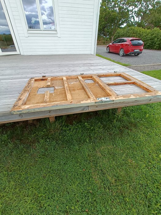 Demonterad träram från en källardörr ligger på en altan, med ett hus och en röd bil i bakgrunden.