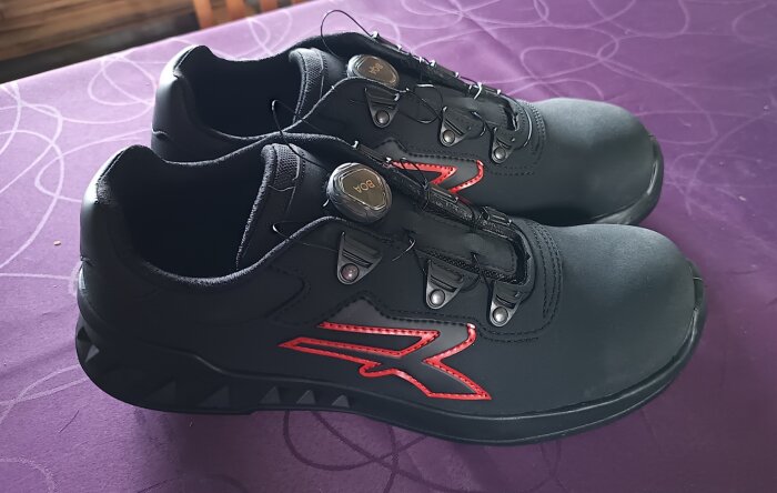 Svarta skyddsskor med röd detalj, snörningssystem från BOA, på lila underlag.