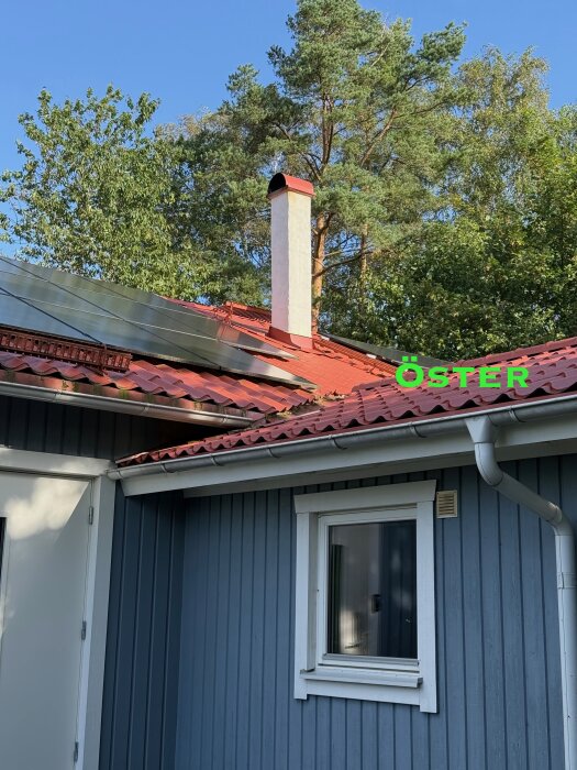 Rött plåttak med solpaneler och murstock, omgiven av skog. Öster markerat i grönt. Problem med läckage nära murstocken.