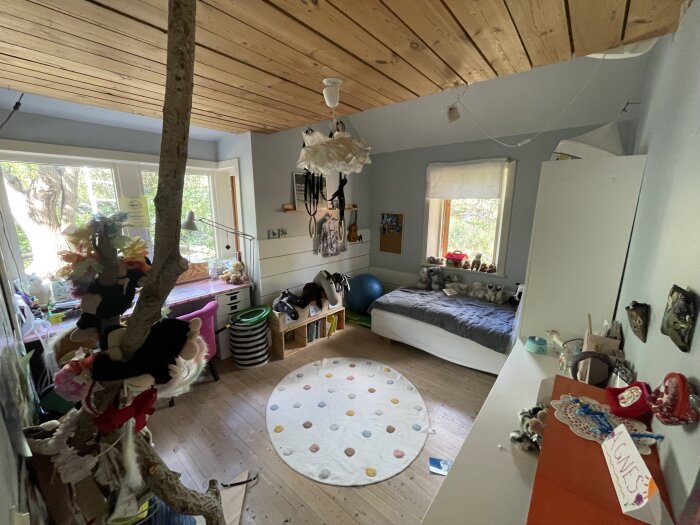 Barnrum med träpaneltak, en säng, skrivbord vid fönstret och ett trädgren som klätterställning. Lekfull inredning med gosedjur och färgglada dekorationer.