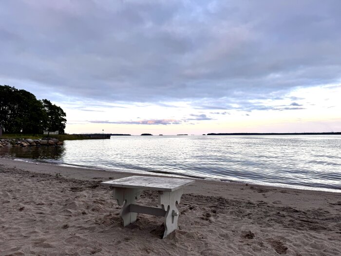 Strand med sand, ett träbord och en molnig himmel vid en stilla sjö med en liten brygga och träd i bakgrunden.