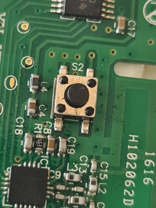 Elektronikkort med en liten rektangulär brytare mitt på kortet, omgiven av kretskomponenter och märkningar.