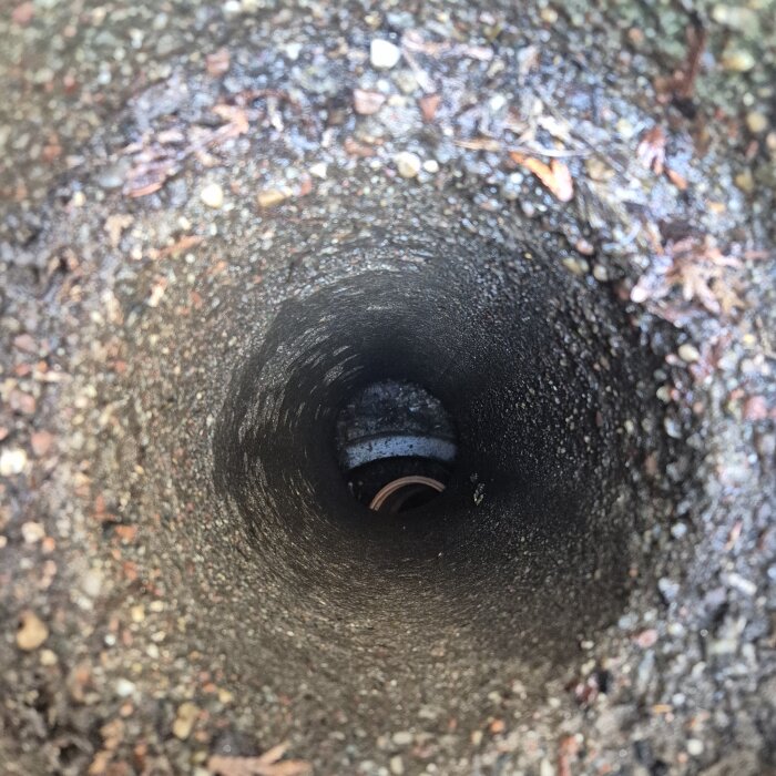 Vy ner i en brunn med snett plaströr som fångar upp regnvatten, sedd från garage med platt tak. Omgivande jord och grus syns i brunnen.