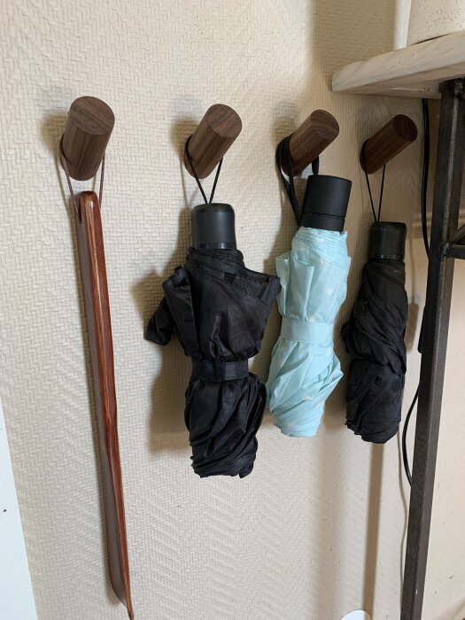 Tre hopfällda paraplyer och en skohorn hänger på träkrokar på en ljus vägg.
