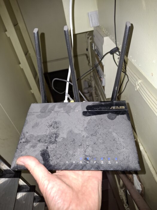 En svart router hålls i en hand, placerad i en källartrappa. Routern har tre antenner och är dammig.
