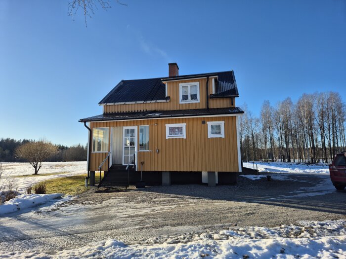 Gult Per Albin torp med svart tak, verandra och snö runtomkring, omringat av träd och åkermark, solig vinterdag.