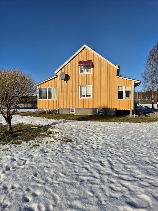 Gult Per Albin torp med sadeltak, snö på marken och klarblå himmel. Trädet i förgrunden och tillbyggd sektion på huset.