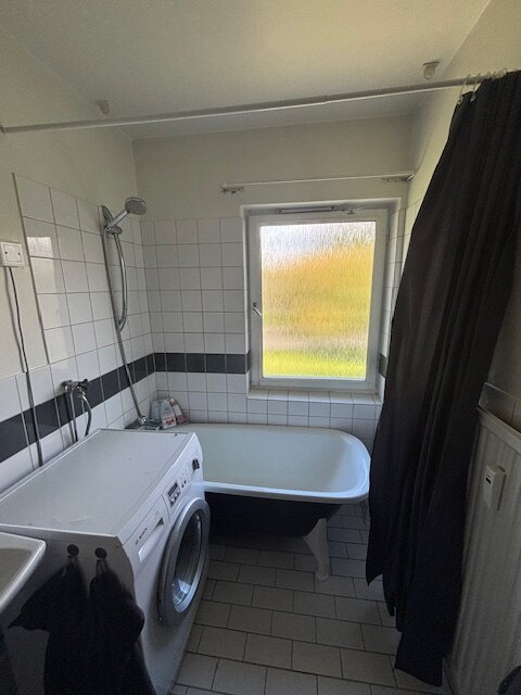 Badrum med gammalt badkar, vitt kakel, duschhuvud, tvättmaskin, svart duschdraperi och fönster med frostat glas.