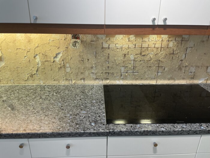 Skadad köksvägg efter borttaget kakel ovanför granitbänkskiva och spishäll, området synligt mellan vita köksskåp.