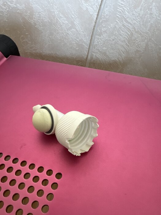 En vit plastdel, trasig med sprickor, märkt "Dispenso Patented Made in SWEDEN", ligger på en rosa perforerad yta.
