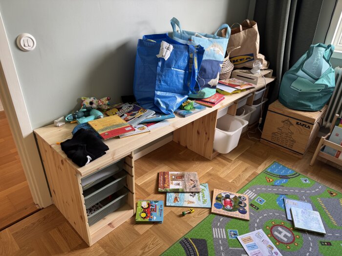 Barnrum med hemmabyggt skrivbord fullt med böcker och leksaker, IKEA-kassar och en lekmatta med bilvägsmönster syns på golvet.