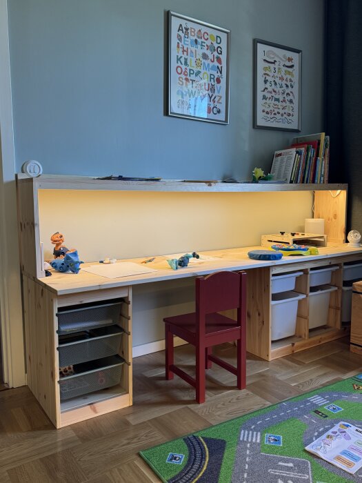 Träskrivbord med hyllplan och inbyggd belysning, barnstol framför. Böcker och leksaker på bänken, affischer på väggen ovanför.