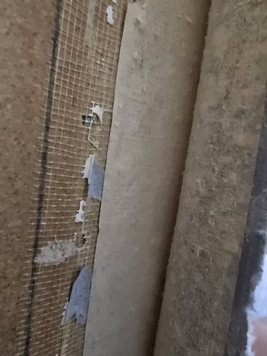 Vägg med borttagen vävtapet, avslöjar rå yta och nätstruktur.