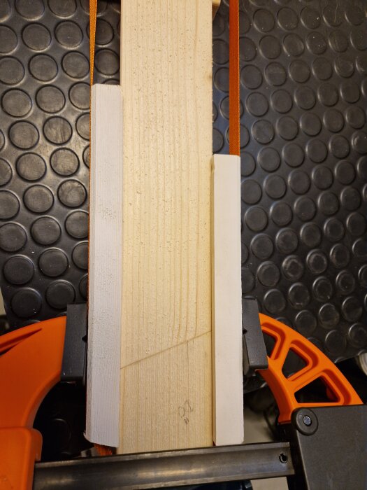 Träbräda med sned skarv, klämd i tving med oranget handtag, liggande på svart mönstrat underlag.