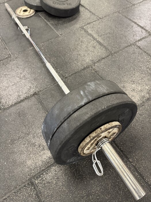Skivstång med viktskivor placerad på ett gymgolv av svart gummi. Ytterligare viktskivor syns i bakgrunden.