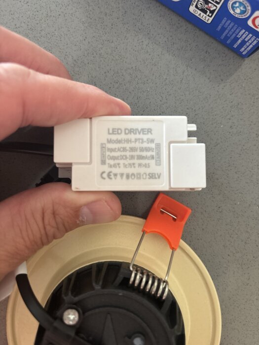 Närbild på en hand som håller en LED-drivare modell HH-PT3-5W med specifikationer synliga. En spotlightdel med orange klämma syns också.