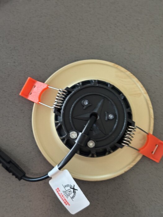 Rund spotlight med svarta och röda kopplingar samt etikett med text, för installation i tak, fotograferad på närbild.