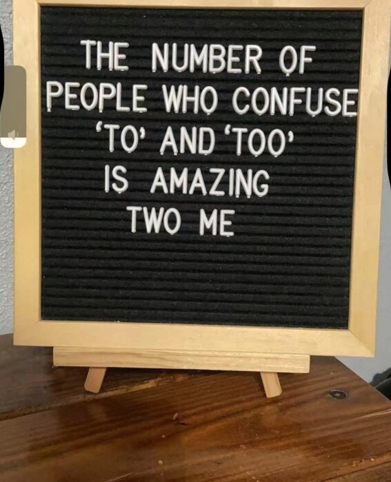 Svart anslagstavla med skämttext: "The number of people who confuse 'to' and 'too' is amazing two me", på ett träbord.